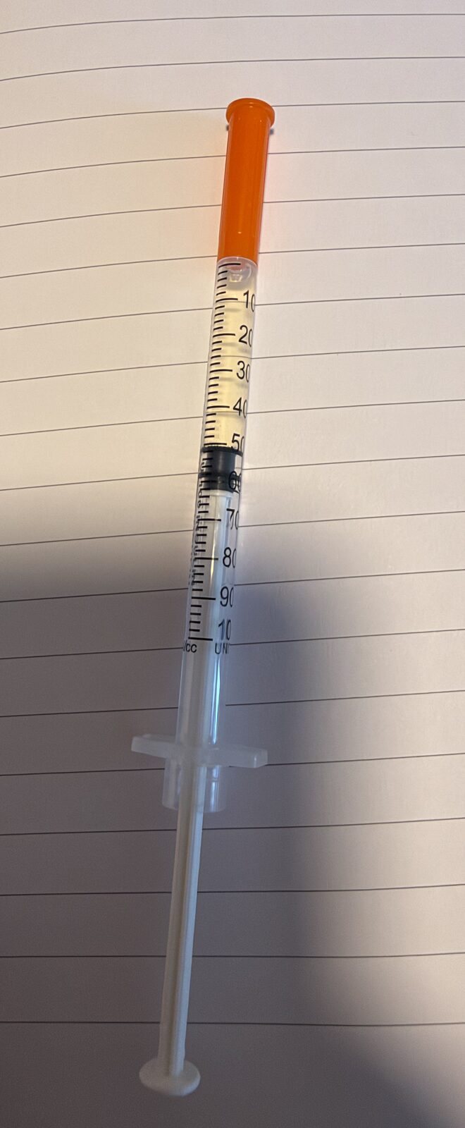 An insulin syringe with an orange cap lying on lined paper, partly filled with a pale yellow liquid