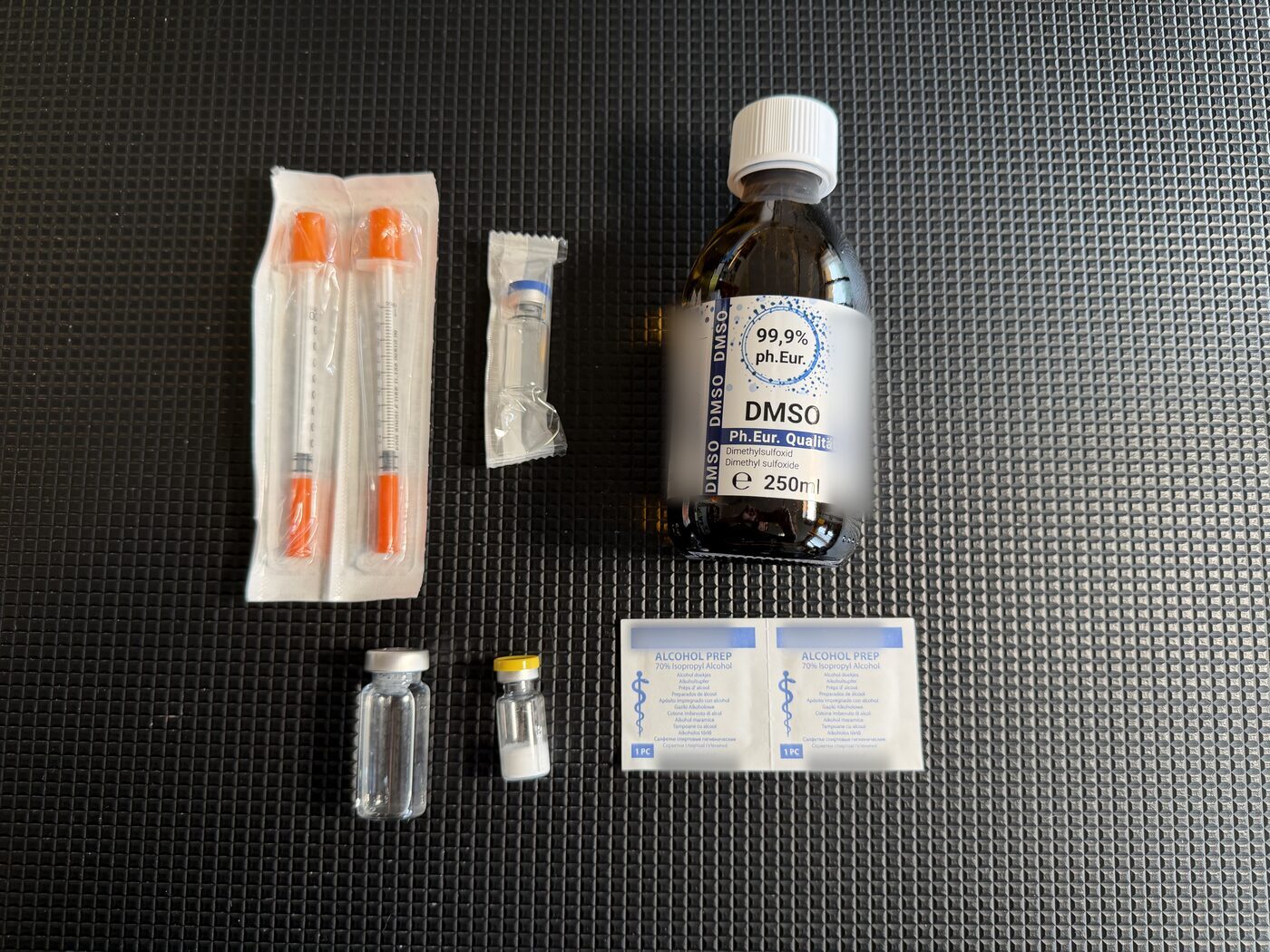 Materiały rozłożone na ciemnej macie: dwie strzykawki insulinowe 1 ml w opakowaniach, butelka DMSO w jakości Ph. Eur., mała fiolka z białym proszkiem, pusta szklana fiolka, fiolka wody bakteriostatycznej jeszcze w opakowaniu i dwa gaziki nasączone alkoholem